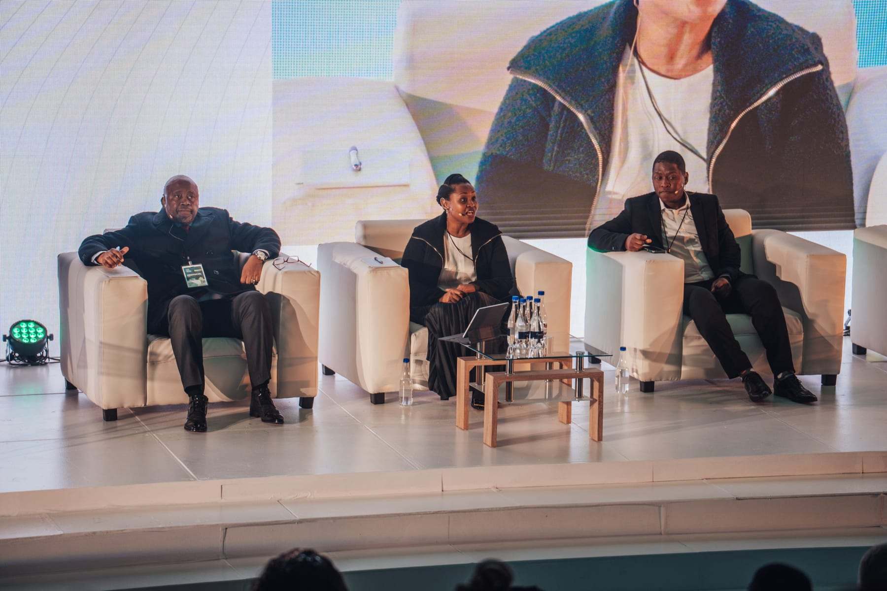 A panel on stage in white chairs, with a speaker on the LED screen behind them