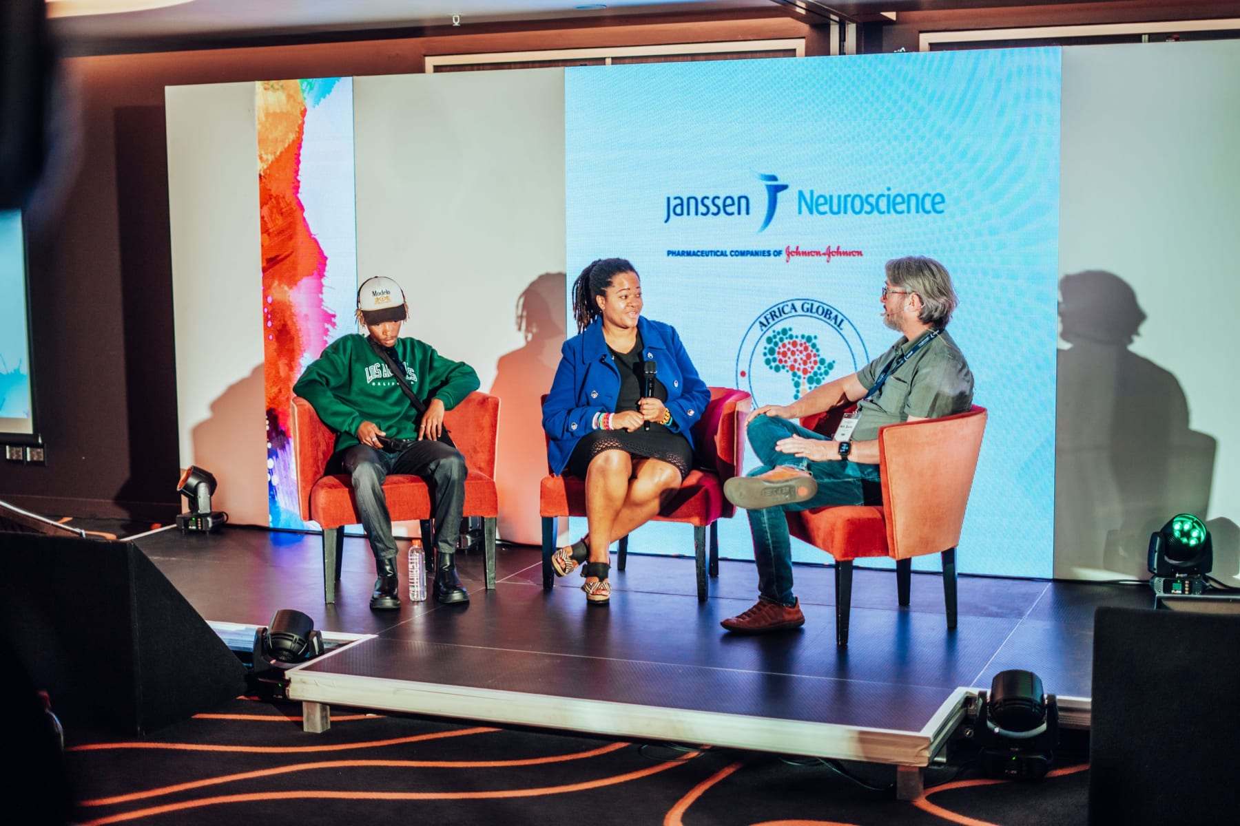 A panel in orange chairs on stage under the screens