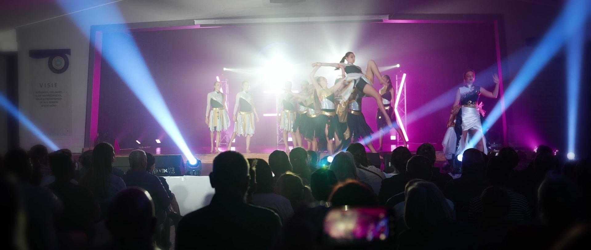 A school show on stage under moving-head beams, seen from the audience