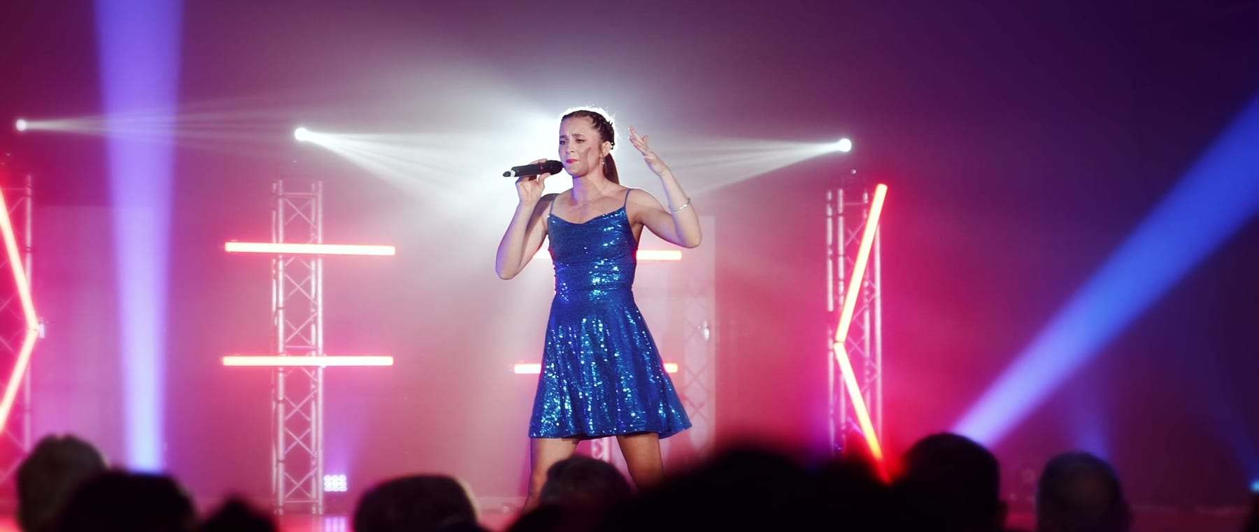 A singer in a blue sequin dress on stage, framed by pink light and beams