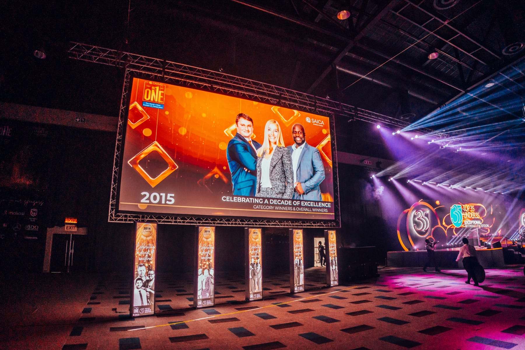 A big screen on stage showing past winners from 2015, with LED towers below it