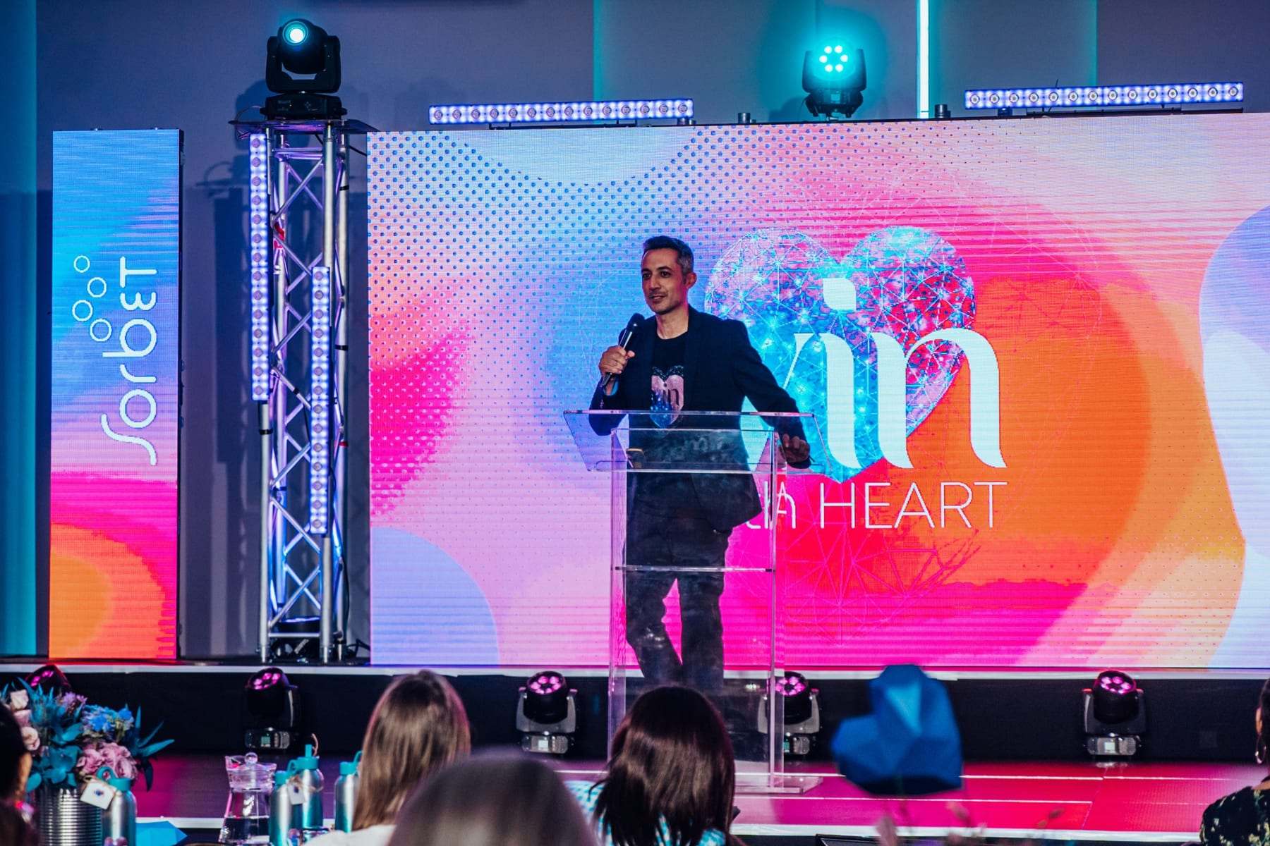 A speaker with a microphone in front of the Win with HEART LED wall